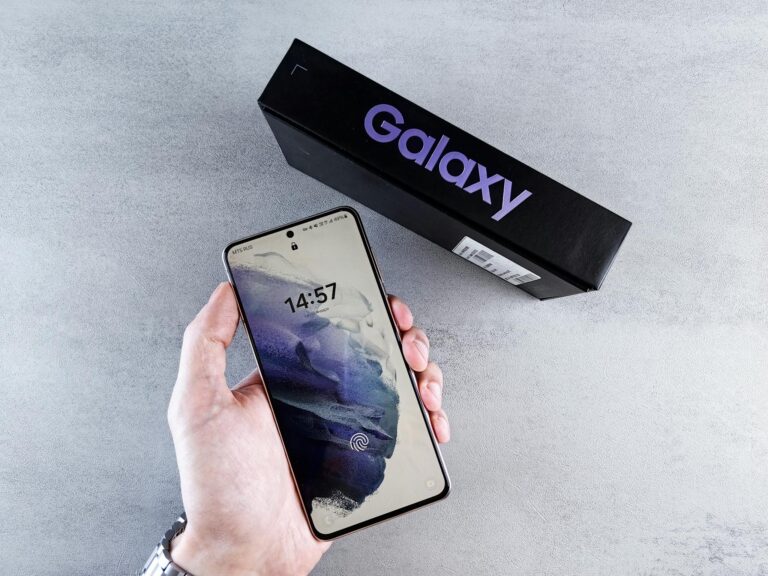 Close-up of a hand holding a smartphone with a Galaxy box on a neutral background.